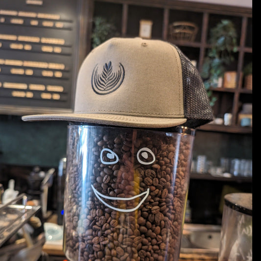 A Throat Punch trucker hat with a rosetta latte art on the front sits on top of a coffee hopper with a smiley face drawn on it. 